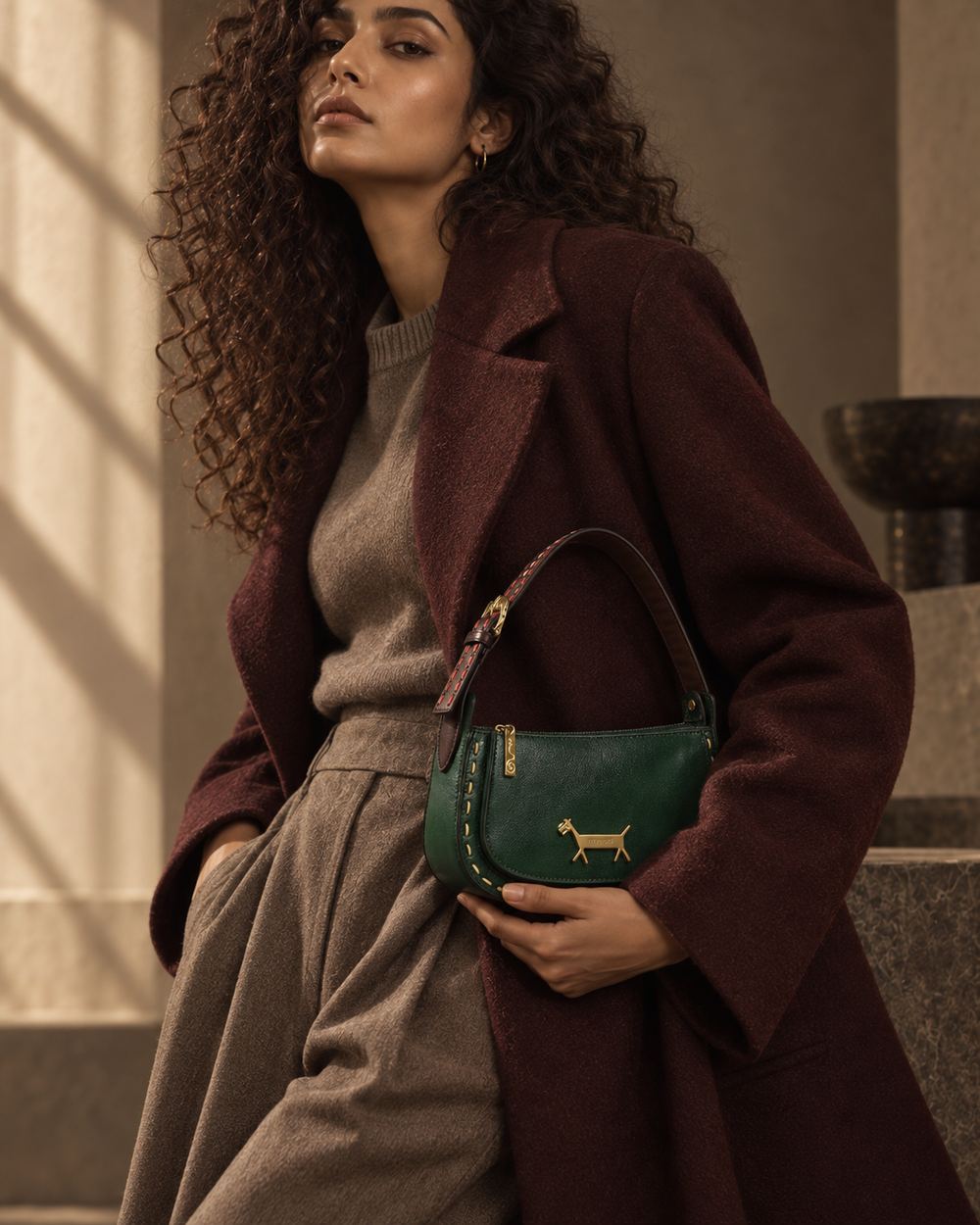 The model holding the green leather bag at her waist, maroon coat over a grey knit and wide trousers.