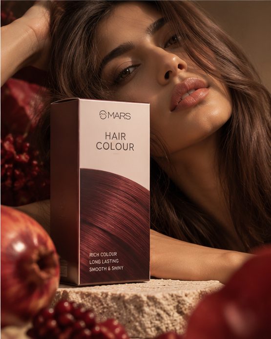 A model resting against stone beside a MARS hair colour carton, pomegranates split open in the warm light around her.