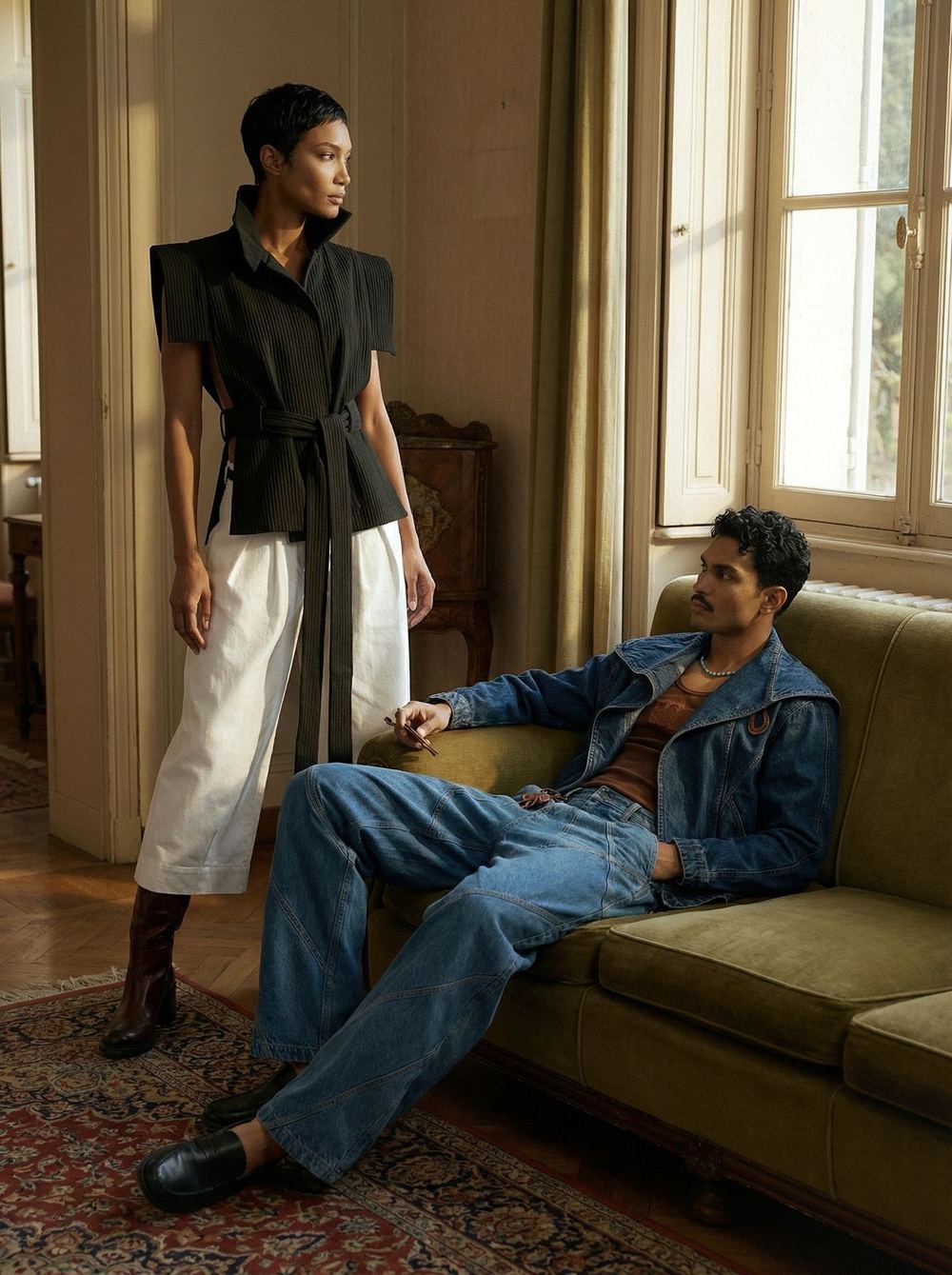 Two models in a sunlit period interior, one standing in a pleated black jacket and white trousers, one seated in denim on a green velvet sofa.