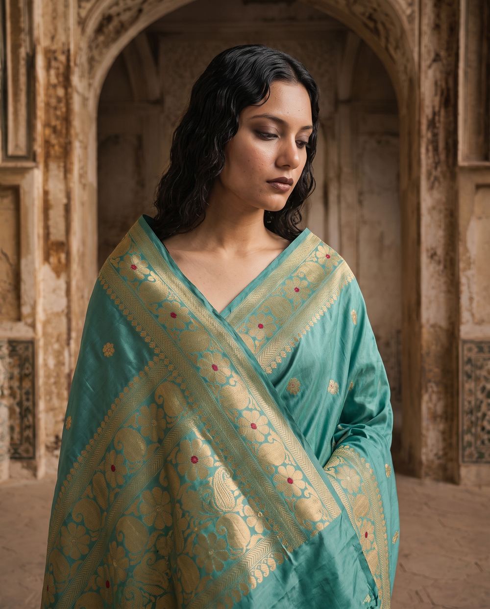 Teal Banarasi silk saree with gold zari, sandstone arch behind.