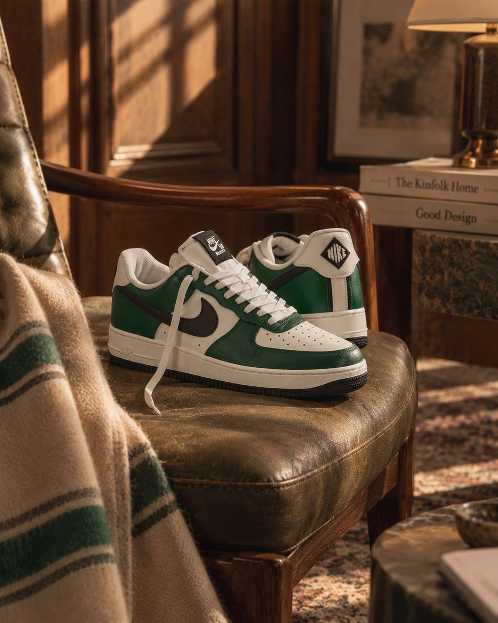 The sneakers on an olive leather chair seat, a folded blanket and stacked books alongside in warm window light.