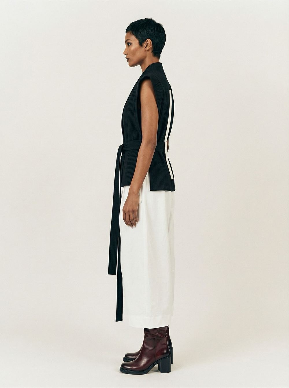 Side profile of a black structured vest over a white column skirt with oxblood boots.